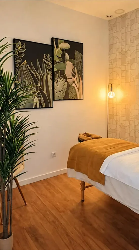 Sala de massagens com marquesa coberta por manta mostarda, quadros botânicos e candeeiro de luz quente